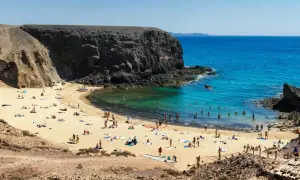 Plaża z turystami kąpiącymi się w turkusowej wodzie, otoczona skalistymi klifami.
