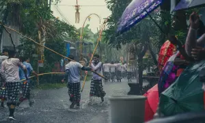 Deszczowa procesja na Bali, kiedy jest sezon na bali. Mężczyźni w tradycyjnych strojach niosą drewniane tyczki, a widzowie chronią się pod kolorowymi parasolami.