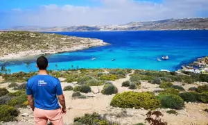 Widok na turkusowe morze i skaliste wybrzeże. Mężczyzna w koszulce "Glob troper" podziwia krajobraz. Tak, Malta jest w Unii Europejskiej.