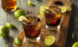 Dwa drinki z colą i lodem, ozdobione ćwiartkami limonki. W tle butelka z bursztynowym płynem. To klasyczny rum z colą, czyli co to jest ron.