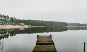 Drewniane molo nad spokojnym jeziorem, z lasem i domkami w tle. To widok, który może skłonić do zastanowienia, ile jest jezior w Olsztynie.