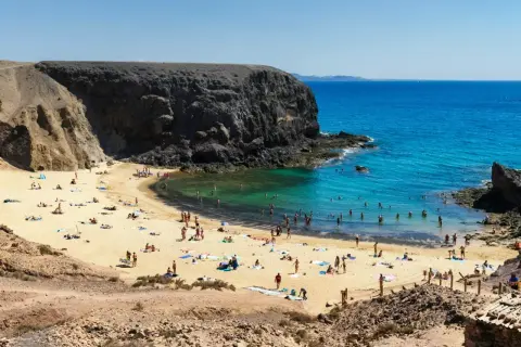 Plaża z turystami kąpiącymi się w turkusowej wodzie, otoczona skalistymi klifami.