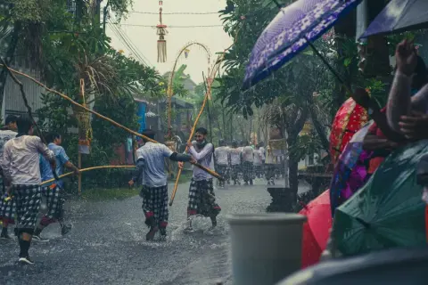 Deszczowa procesja na Bali, kiedy jest sezon na bali. Mężczyźni w tradycyjnych strojach niosą drewniane tyczki, a widzowie chronią się pod kolorowymi parasolami.
