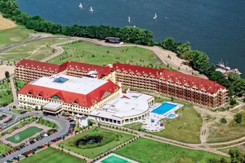 Widok z lotu ptaka na rozległy kompleks hotelowy z czerwonymi dachami, basenem i jeziorem. Czy to pierwszy hotel Gołębiowski?