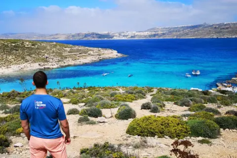 Widok na turkusowe morze i skaliste wybrzeże. Mężczyzna w koszulce "Glob troper" podziwia krajobraz. Tak, Malta jest w Unii Europejskiej.