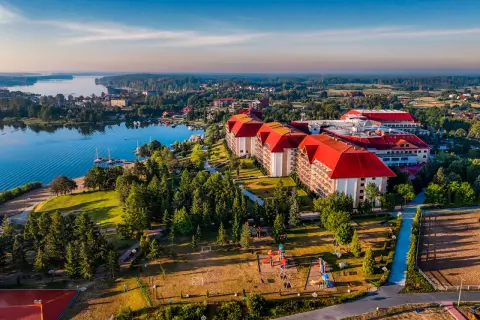 Hotel Gołębiewski w Mikołajkach, z czerwonymi dachami, otoczony zielenią i jeziorem, wygląda jak miejsce z bajki.