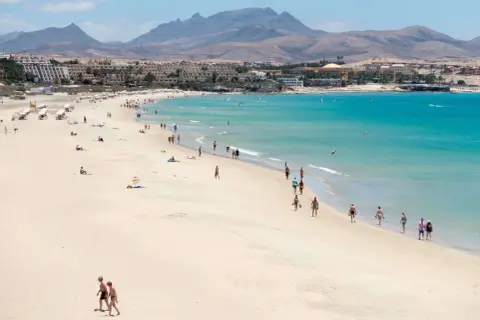 Słoneczna plaża z turystami, turkusowe morze i góry w tle. Czy lot na Wyspy Kanaryjskie jest bezpieczny? Tak, to popularny i bezpieczny kierunek wakacyjny.