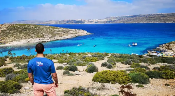 Widok na turkusowe morze i skaliste wybrzeże. Mężczyzna w koszulce "Glob troper" podziwia krajobraz. Tak, Malta jest w Unii Europejskiej.