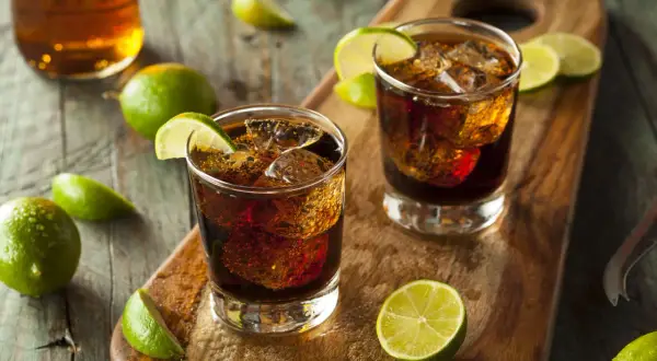Dwa drinki z colą i lodem, ozdobione ćwiartkami limonki. W tle butelka z bursztynowym płynem. To klasyczny rum z colą, czyli co to jest ron.