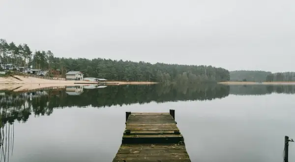 Drewniane molo nad spokojnym jeziorem, z lasem i domkami w tle. To widok, który może skłonić do zastanowienia, ile jest jezior w Olsztynie.