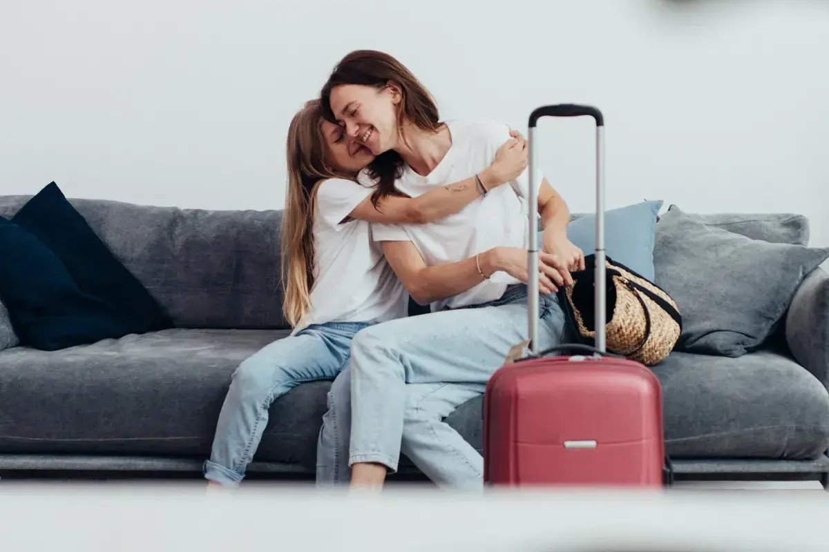 Mama i córka przytulają się na kanapie obok walizki. Wszyscy, którzy chcą podróżować, mogą zameldować się w hotelu.