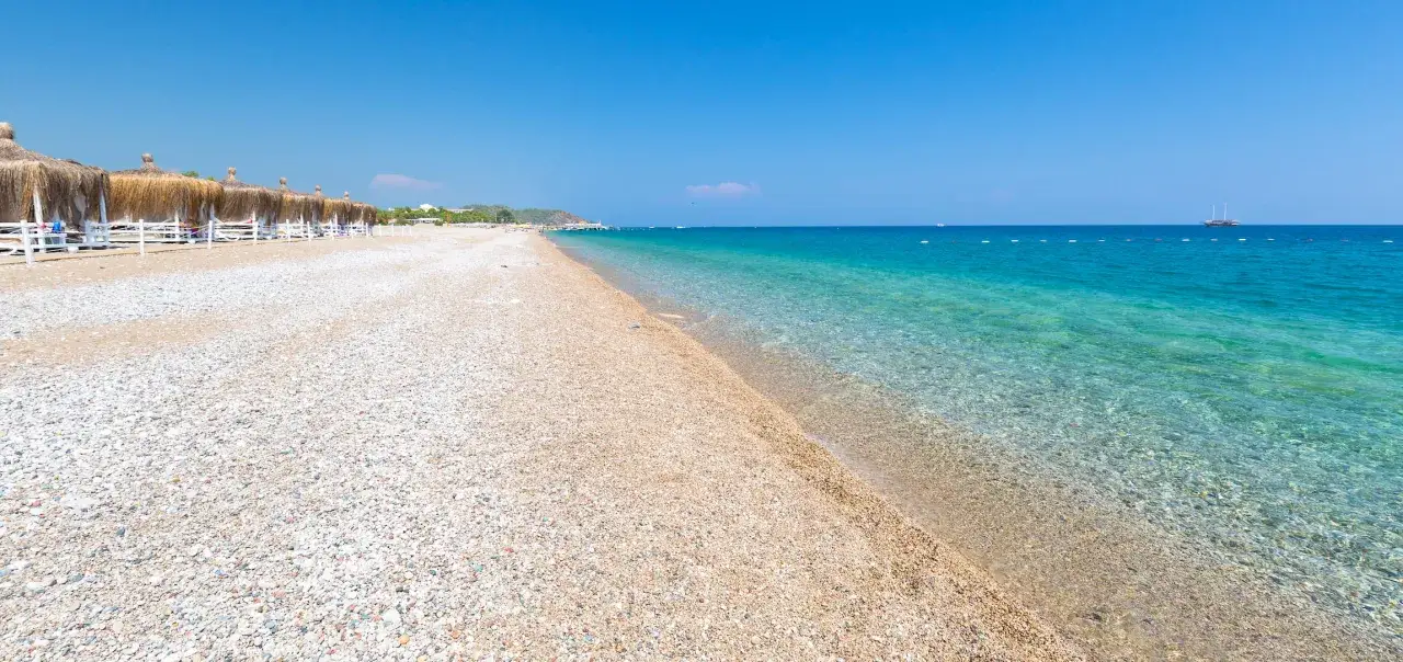 Piękna, kamienista plaża w Antalyi z turkusową wodą i zacienionymi leżakami. Czy Antalya jest bezpieczna? Tak, to popularny i bezpieczny kurort.