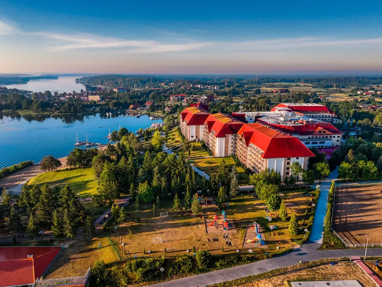 Widok z lotu ptaka na kompleks hotelowy z czerwonymi dachami, placem zabaw i jeziorem. Czy to pierwszy hotel Gołębiowski?