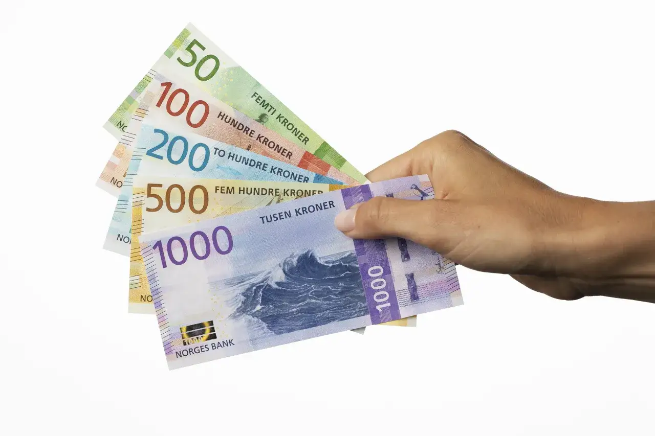 Dłoń trzyma banknoty: 50, 100, 200, 500 i 1000. To norweskie korony (NOK).