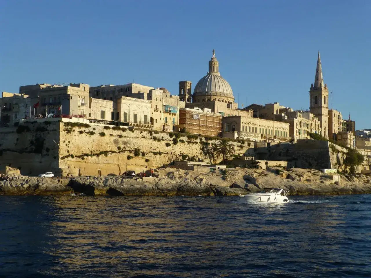 Malta, kiedy jest najcieplej, wita łodzią motorową na tle zabytkowego miasta z kopułą i wieżą kościelną.