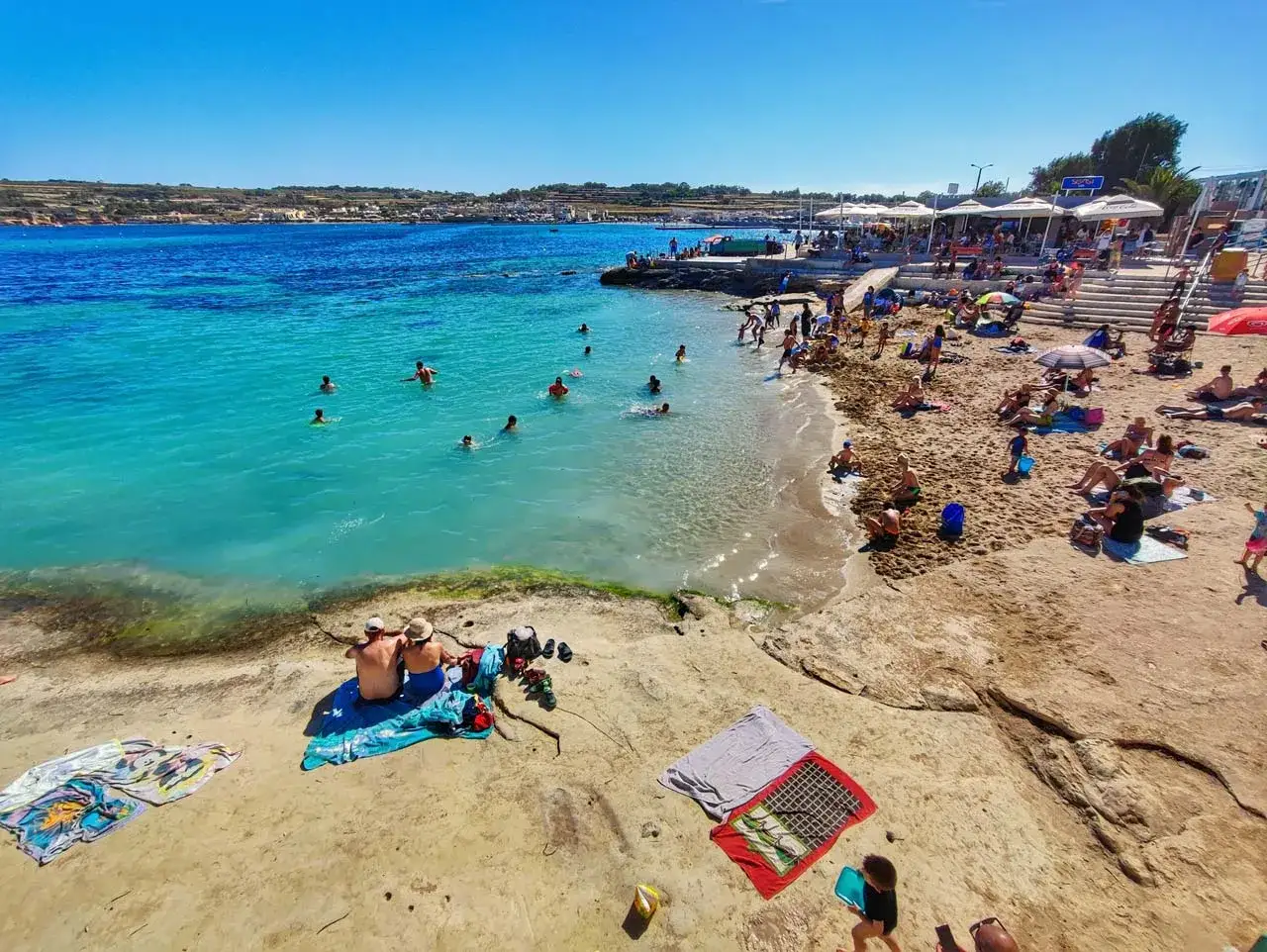 Słoneczna plaża z turkusową wodą, ludzie kąpią się i opalają. Idealne miejsce, by sprawdzić, gdzie cieplej w kwietniu: Cypr czy Malta.