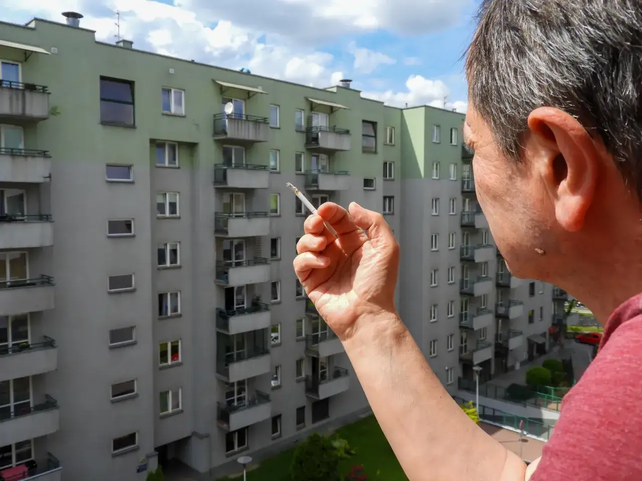 Mężczyzna z papierosem na balkonie, zastanawia się, czy w hotelach można palić na balkonie. W tle blok mieszkalny.