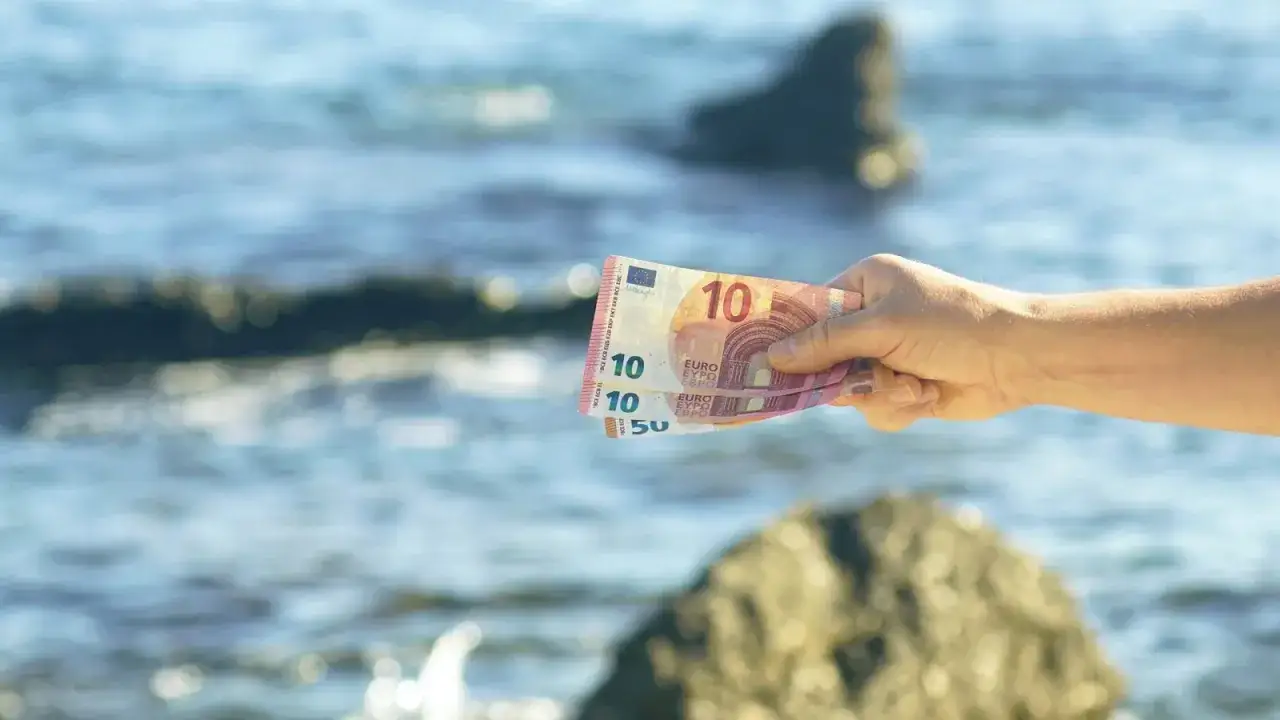Dłoń trzyma banknoty euro, sugerując, jaką walutę ma Czarnogóra. W tle widać morze i skały.