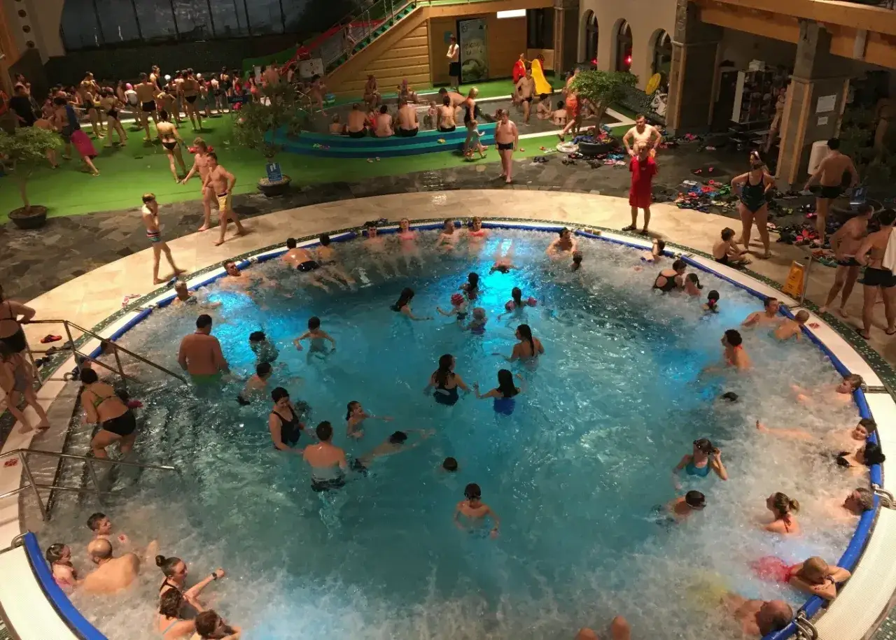Tłum ludzi w basenie z hydromasażem. Czy termy chochołowskie są naturalne? Woda jest ciepła i błękitna, a wokół basenu widać zielone otoczenie.