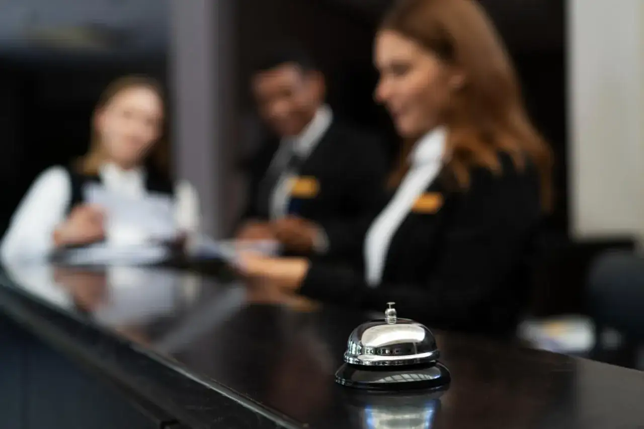 Recepcjonistka w hotelu obsługuje gości przy ladzie. W tle widać innych pracowników.
