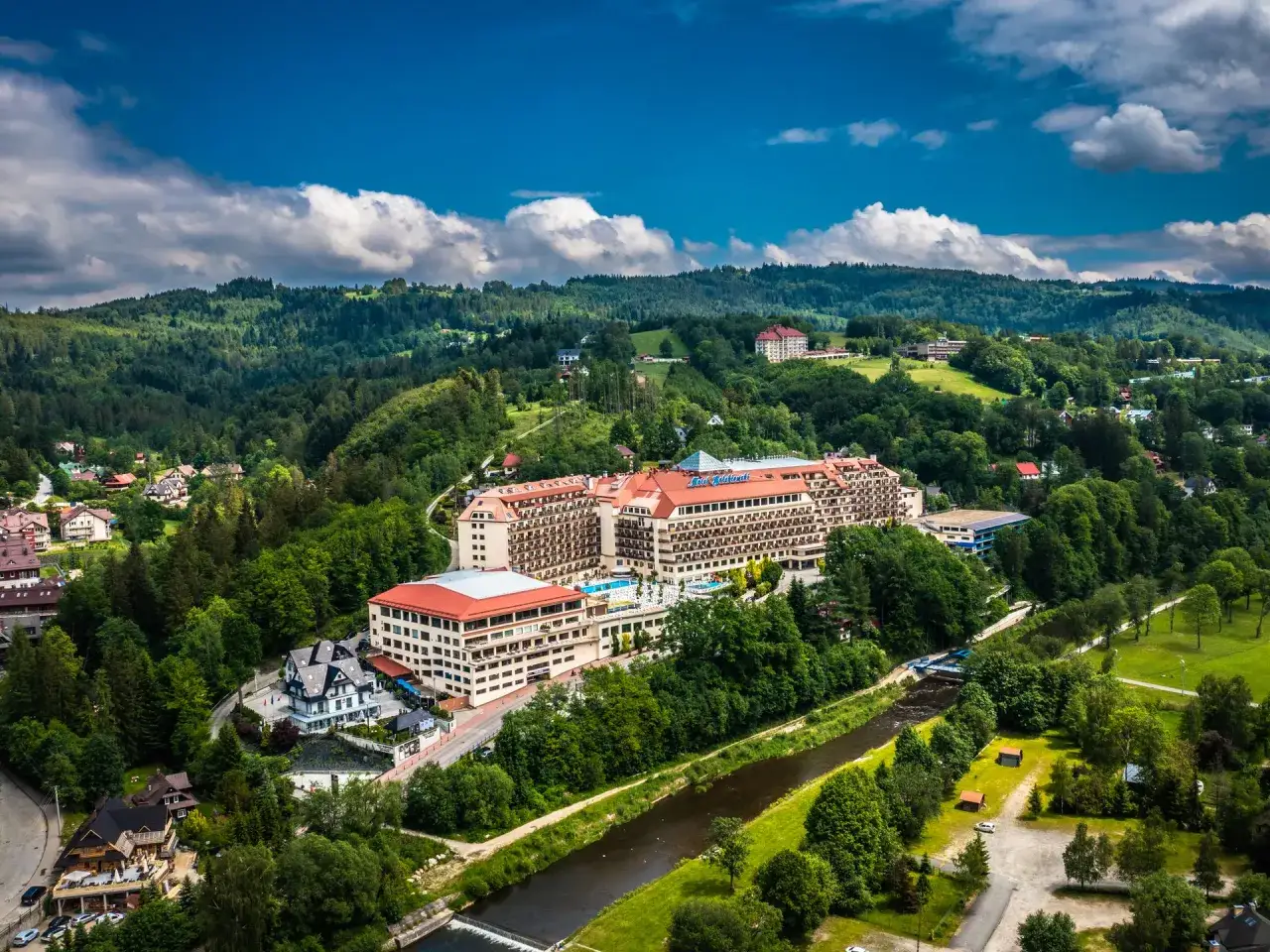Widok z lotu ptaka na Hotel Gołębiewski w otoczeniu zielonych wzgórz i rzeki. To jeden z wielu hoteli Gołębiewskich w Polsce.