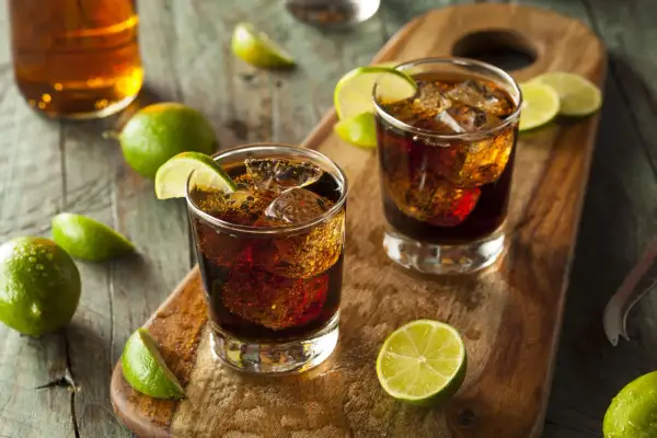 Dwa drinki z colą i lodem, ozdobione ćwiartkami limonki. W tle butelka z bursztynowym płynem. To klasyczny rum z colą, czyli co to jest ron.
