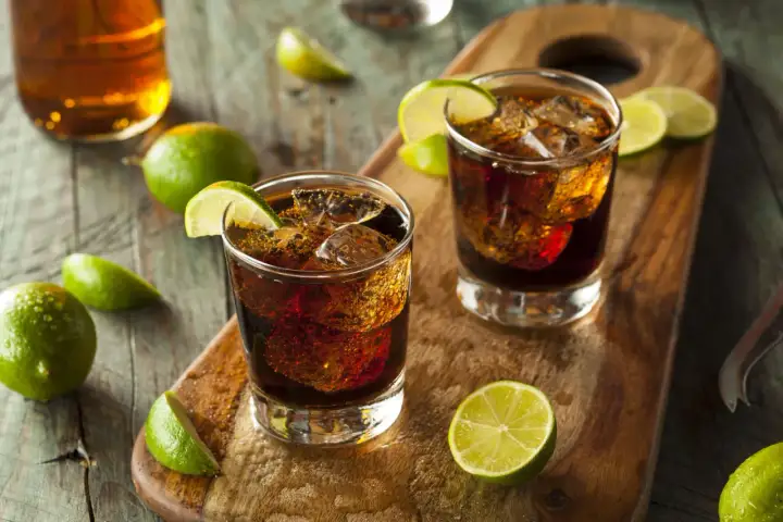 Dwa drinki z colą i lodem, ozdobione ćwiartkami limonki. W tle butelka z bursztynowym płynem. To klasyczny rum z colą, czyli co to jest ron.